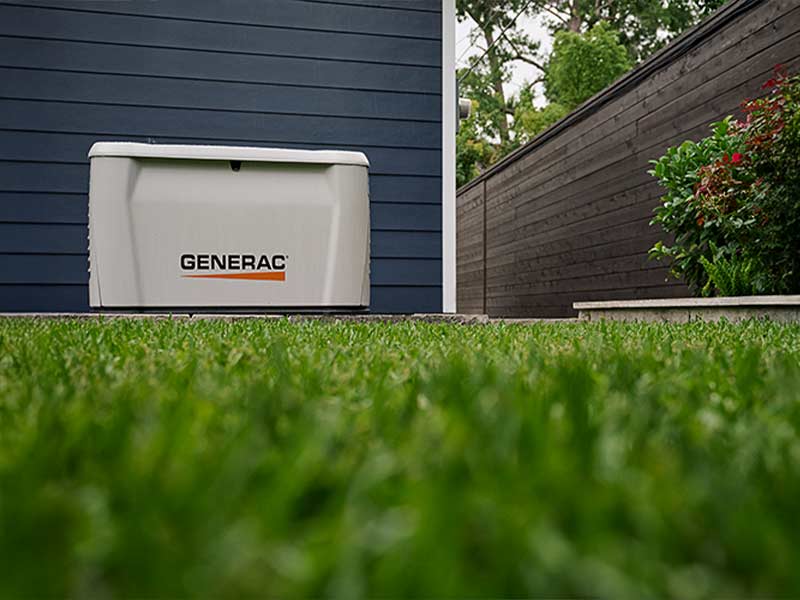 Standby generator on a pad beside a home’s blue siding, with landscaping along the wall